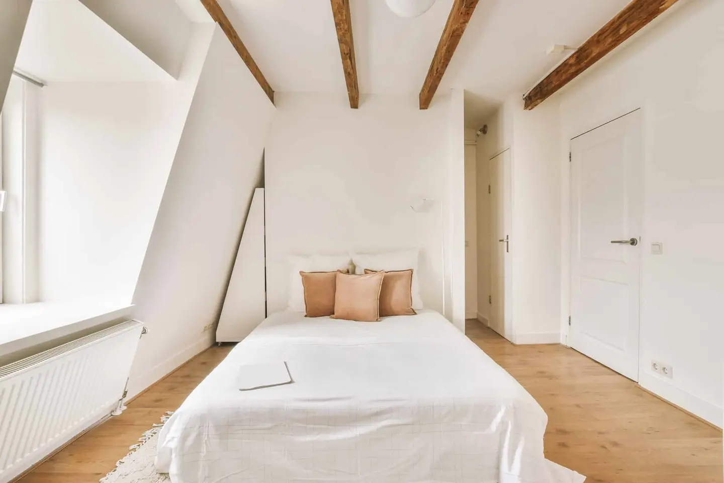 Chambre aménagée sous les combles : murs blancs, poutres apparentes, plafond en pente et parquet clair