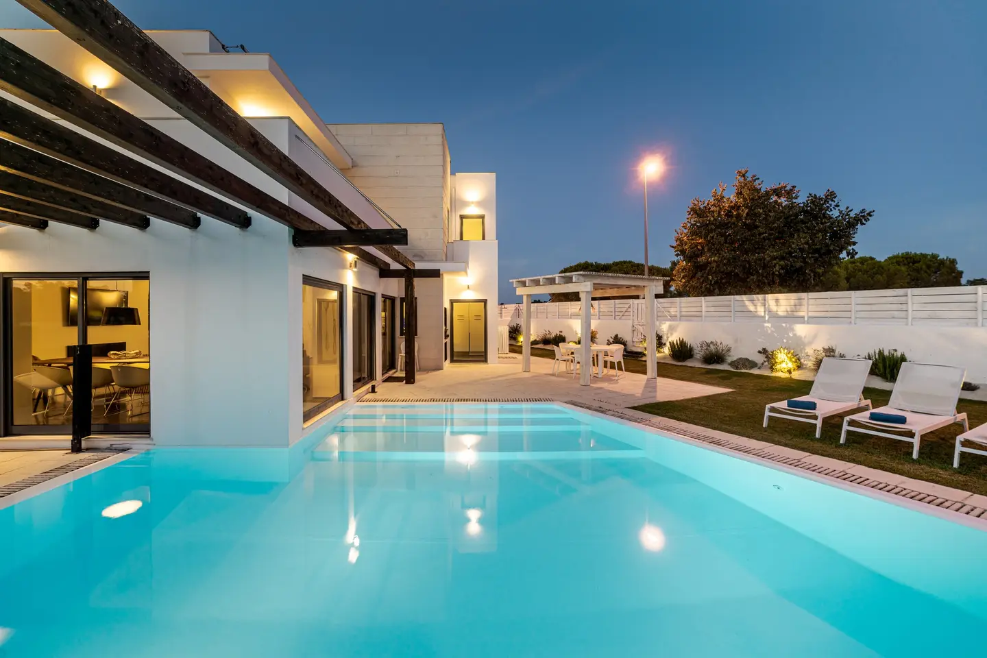 Maison contemporaine au crépuscule : piscine, pergola en bois, pelouse et transats