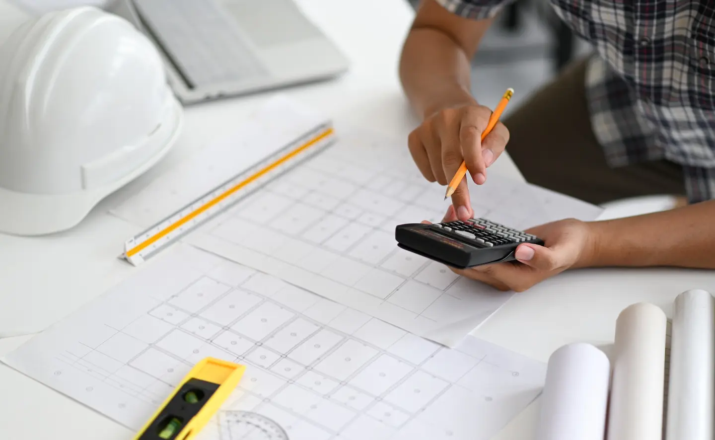 Mains tenant une calculatrice et un crayon au-dessus de plans, à côté d'un casque de chantier