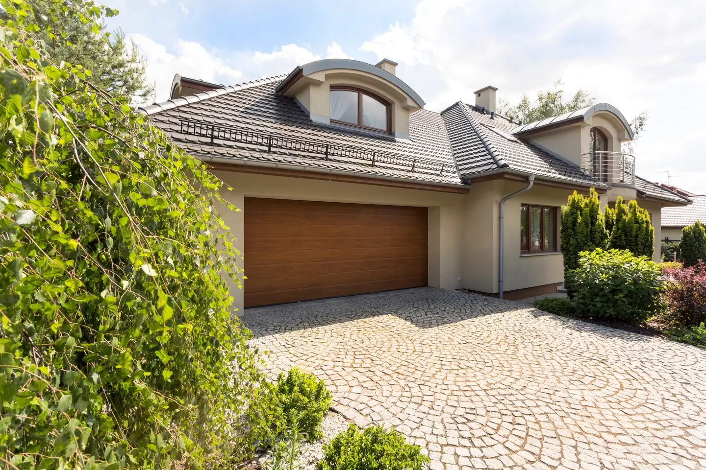 Allée de garage en pavés posés en arcs devant une maison individuelle à porte de garage en bois, bordée d'arbustes
