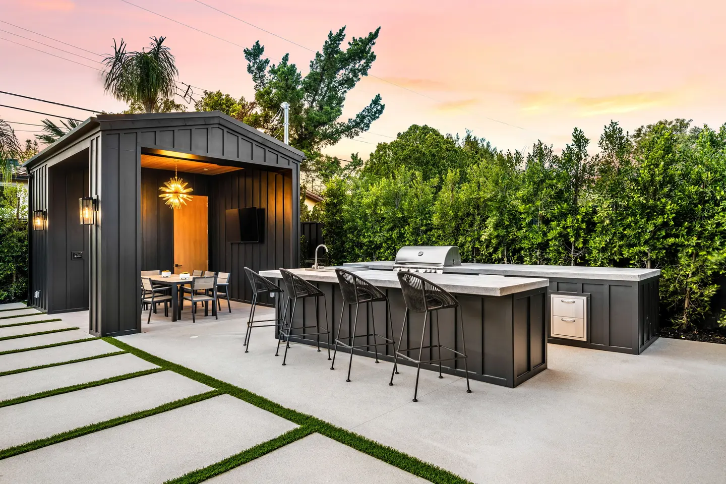 Cuisine d'été au coucher du soleil : îlot avec barbecue encastré, tabourets de bar et abri au bardage noir sur une terrasse en dalles