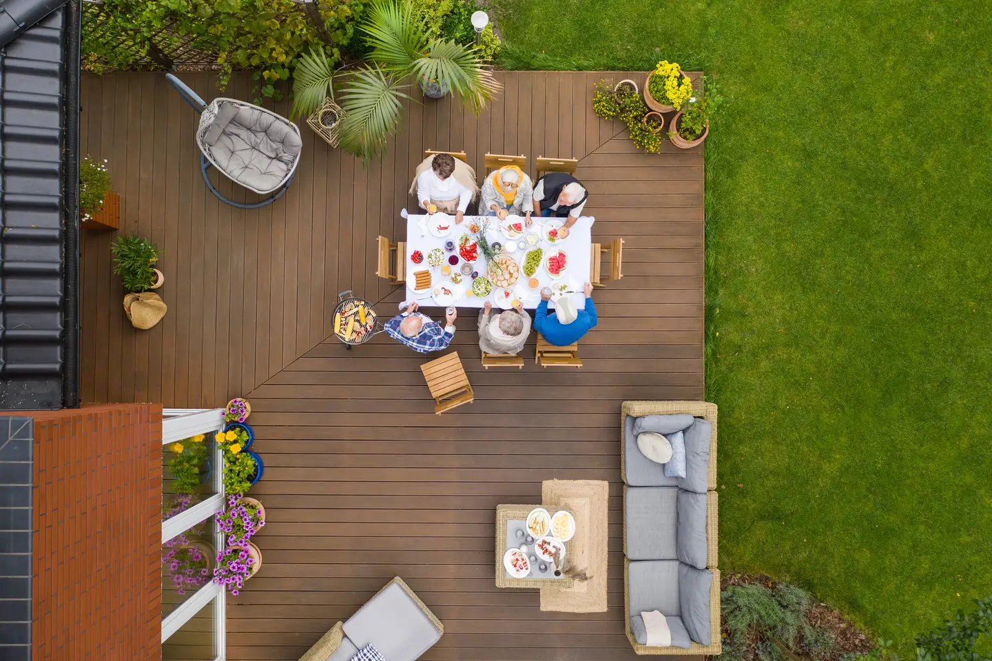 Terrasse en lames de bois vue du dessus, au bord d'une pelouse : une famille à table, un salon de jardin et des plantes en pots