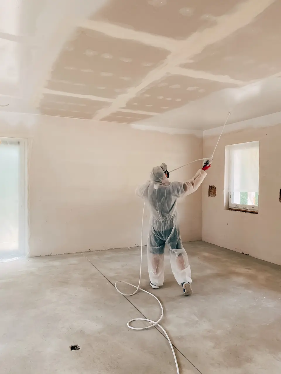 Peintre en combinaison pulvérisant de la peinture au plafond d'une pièce en travaux
