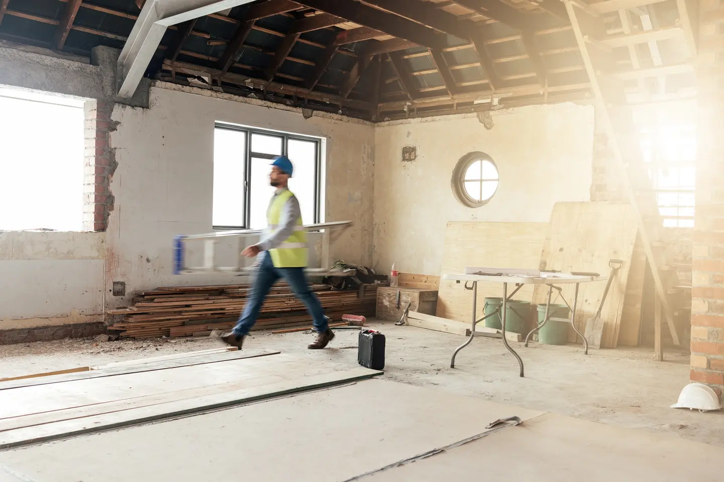 Ouvrier casqué traversant une maison en pleine rénovation : charpente apparente, murs mis à nu et fenêtres