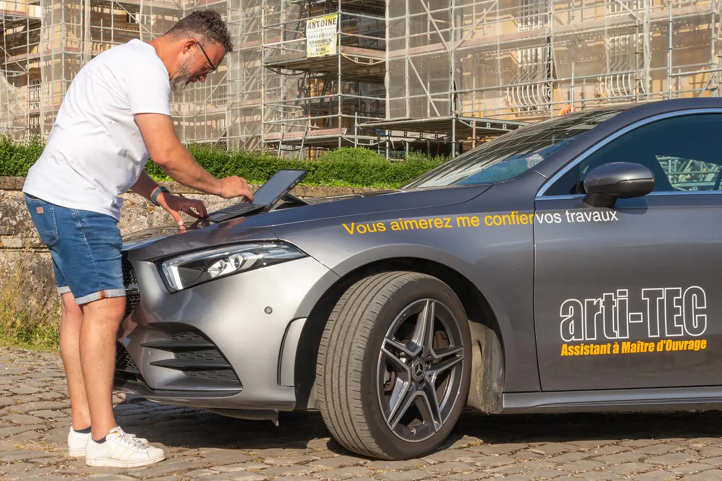 Sébastien Milone consulte sa tablette posée sur le capot de la voiture arti-TEC, devant un immeuble sous échafaudage